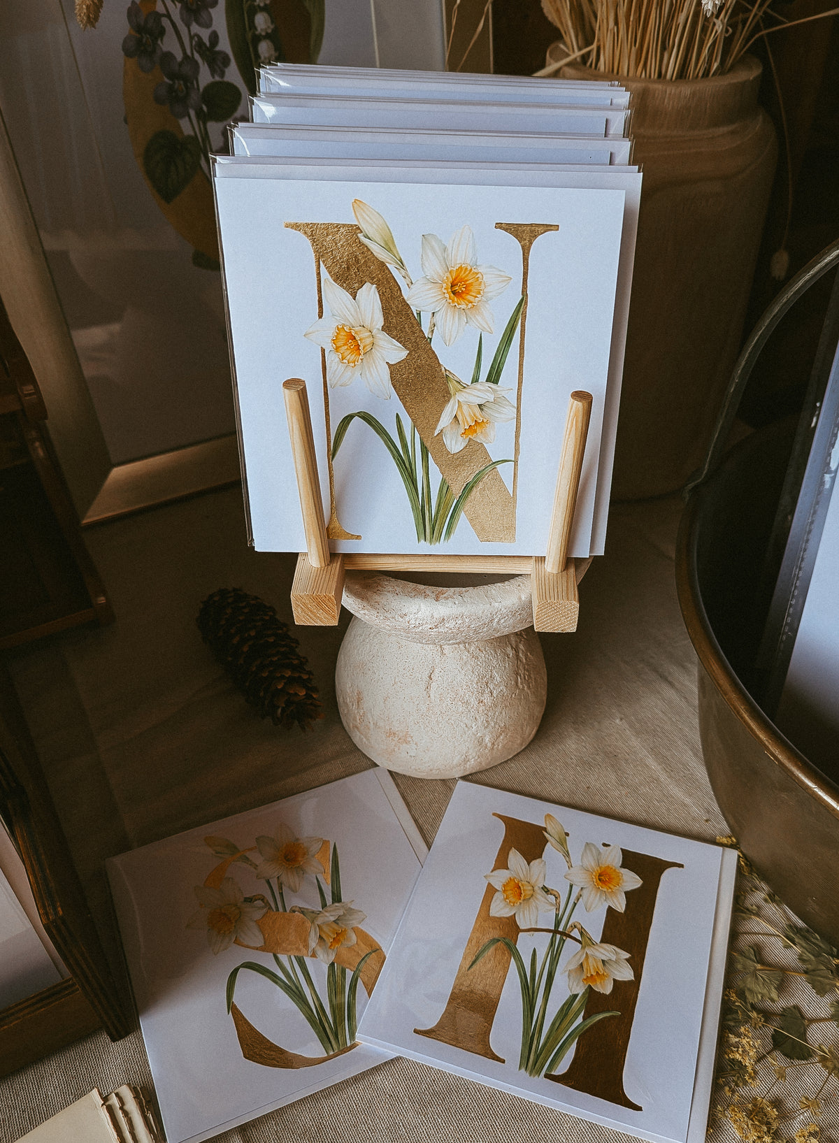 A stack of square art cards displayed on a small wooden stand, featuring a large gold letter 'N' with white narcissus flowers and green stems. Two matching prints lie on the floor in front, with a rustic ceramic pedestal, dried grasses, and warm indoor decor in the background.