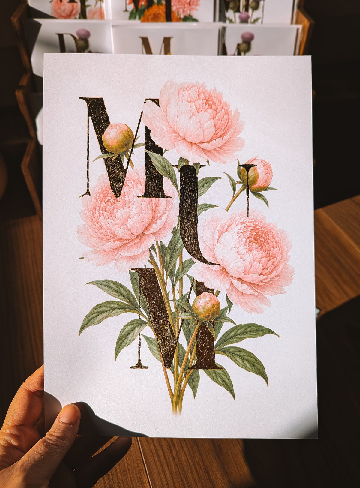 A hand holds a white art print featuring the word 'MUM' in large gold foil serif letters filled with illustrations of pink peonies and green leaves, set against a warm wooden background.