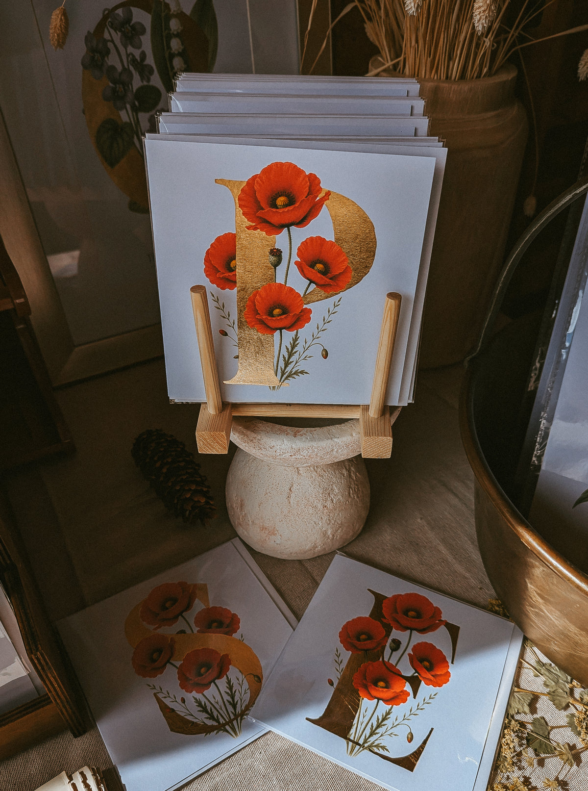 A stack of art cards featuring a large gold letter 'P' decorated with red poppies, displayed on a wooden stand in a rustic indoor setting with dried flowers and a bronze bowl.