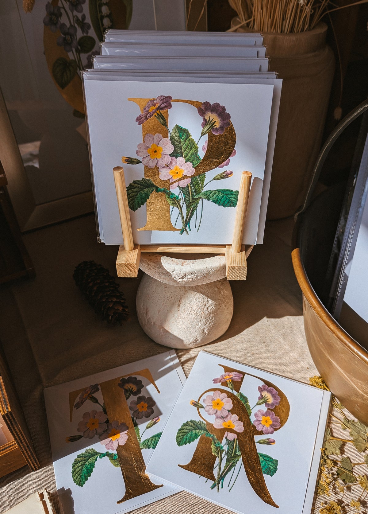 A stack of white art cards featuring decorative gold letter designs with pink and purple primrose flowers and green leaves, displayed on a small wooden easel in a styled indoor setting with a stone pedestal and additional prints on the floor.