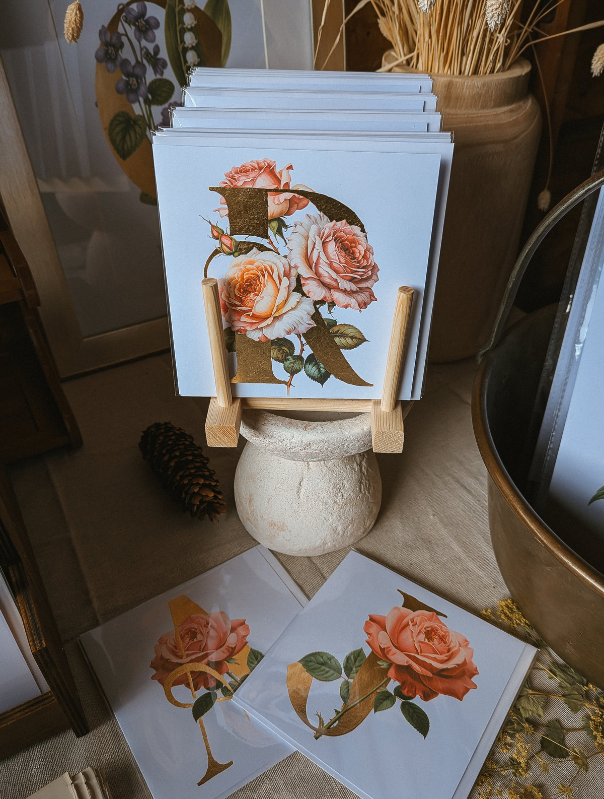 A stack of square art cards displayed on a small wooden stand, featuring a vintage-style rose illustration with a large gold letter 'R'. Additional matching cards are placed on the floor around them in a styled indoor setting with dried flowers and rustic decor.