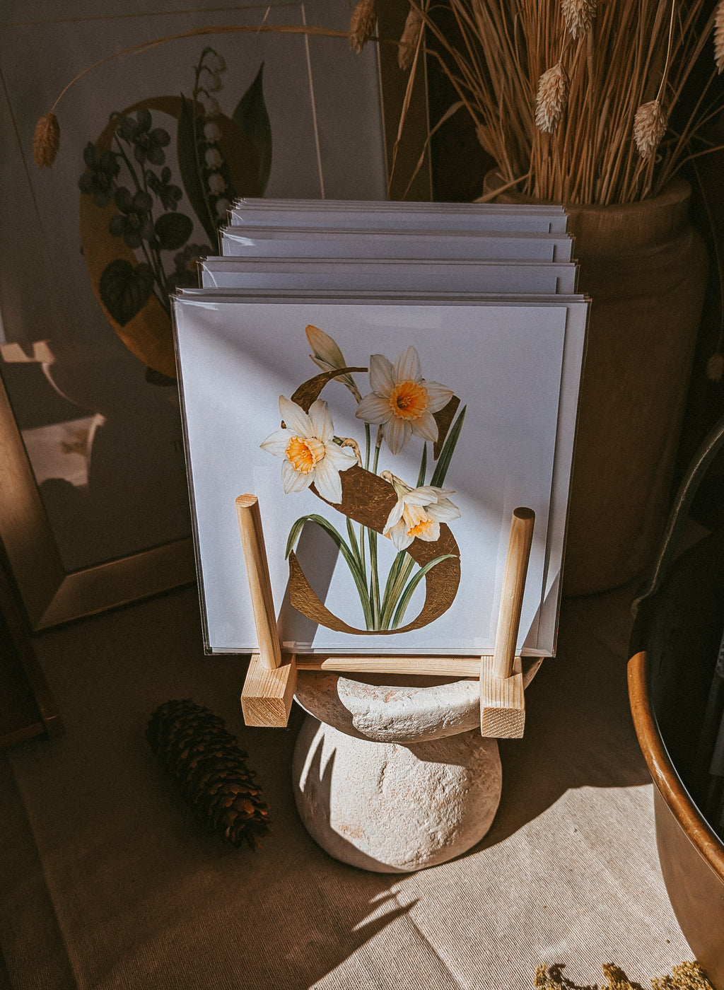 A stack of square cards displayed on a small wooden stand in a styled interior setting. The front print features white narcissus with yellow centers and a large gold letter 'S' on a white background.
