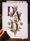 A hand holds a white art print featuring the word 'DAD' in large textured gold foil letters overlaid with detailed illustrations of purple thistles and green leaves, set against a dark indoor background.
