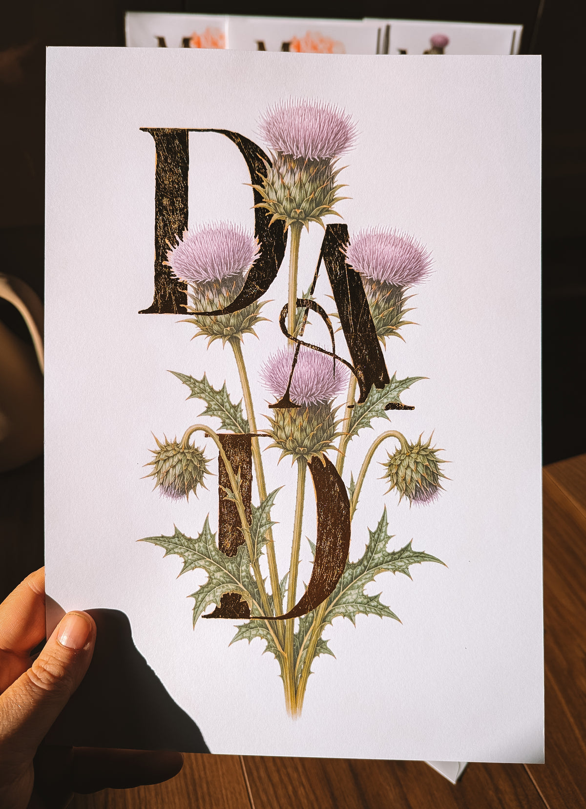 A hand holds a white art print featuring the word 'DAD' in large textured gold foil letters overlaid with detailed illustrations of purple thistles and green leaves, set against a dark indoor background.