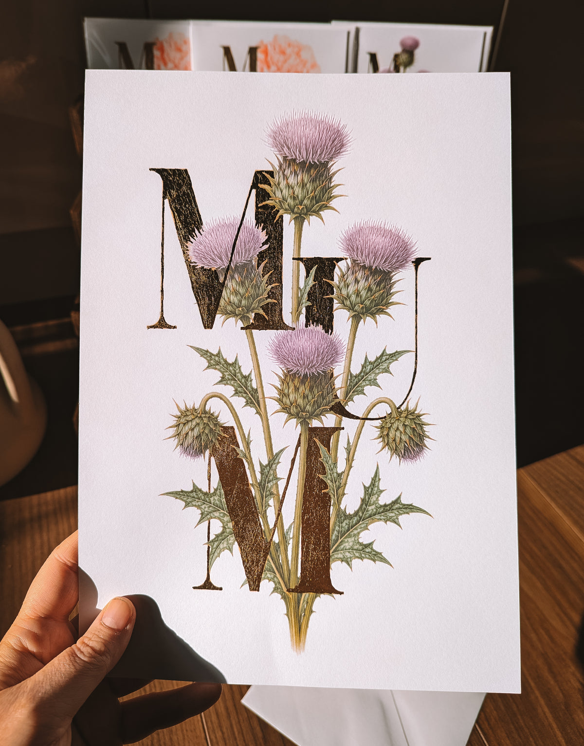A hand holds a white art print featuring the word 'MUM' in large gold foil letters decorated with thistle flowers, on a wooden table.