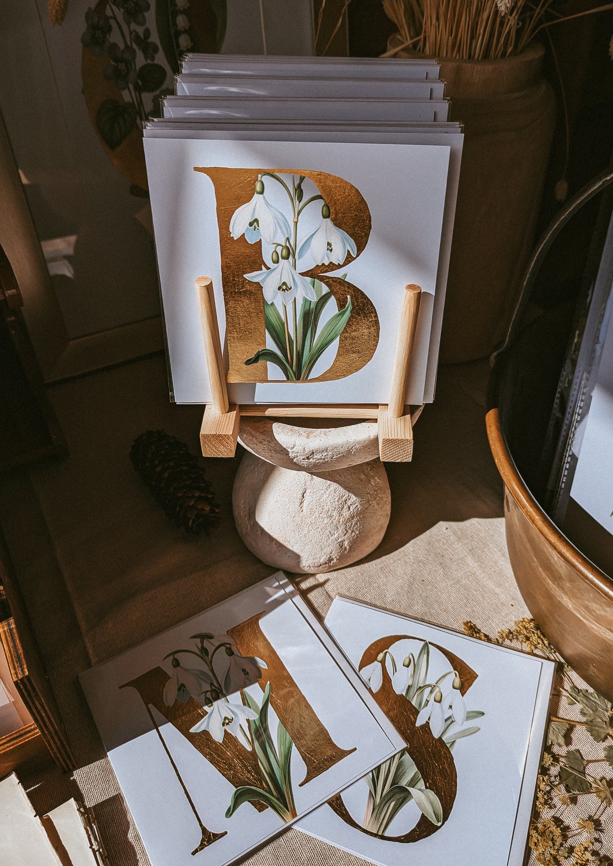 A stack of square art cards displayed on a small wooden stand, featuring large decorative letters with white snowdrop floral illustrations and gold leaf-like accents. Two matching prints lie on the floor in front, set in a rustic styled interior with warm natural light.