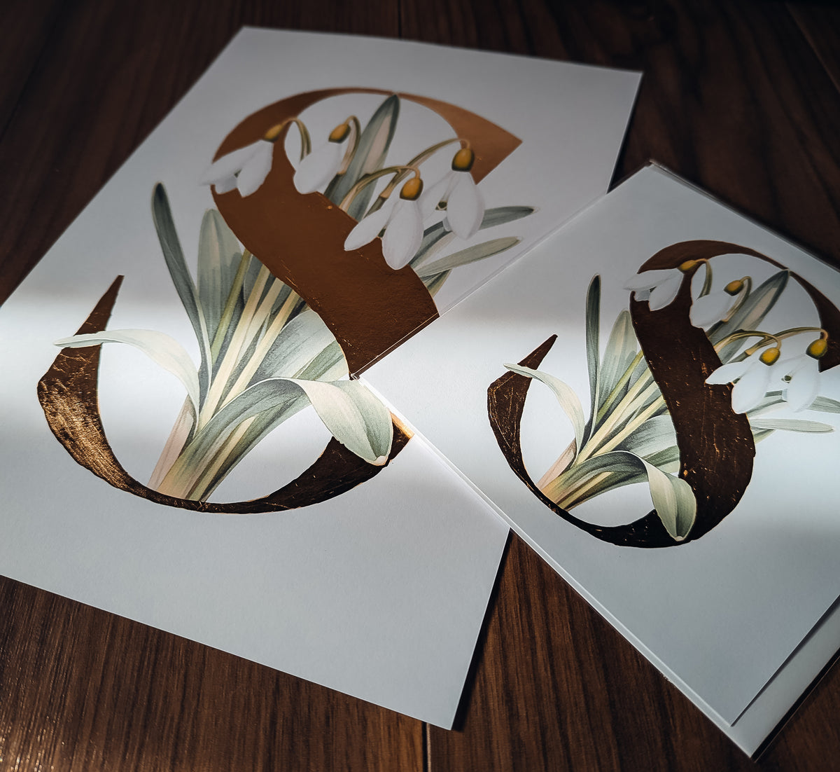 Print & card featuring a large decorative letter 'S' filled with white snowdrop flowers and green leaves, arranged on a dark wooden surface.