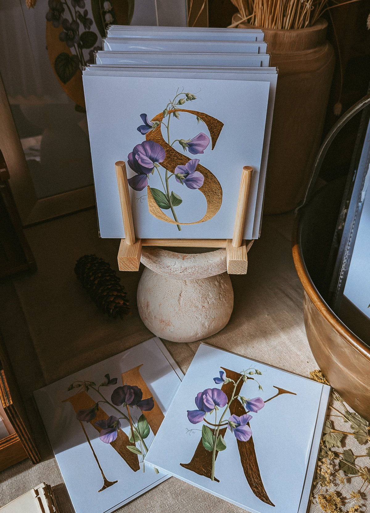A stack of square art cards displayed on a small wooden stand, with two matching prints lying in front on a tabletop. The designs feature large gold letterforms decorated with purple sweet pea flowers and green leaves, set in a warm, rustic interior with a ceramic pot and dried botanicals in the background.