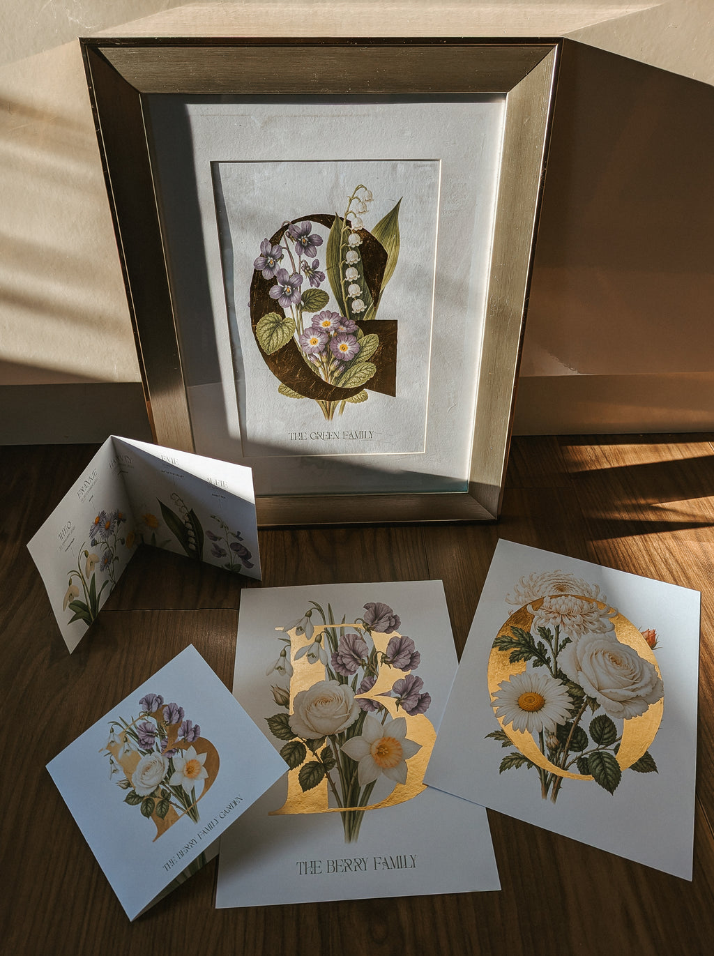 A framed botanical art print and matching printed cards arranged on a wooden surface in warm sunlight. The central framed piece shows a stylised letter 'G' with purple flowers and green leaves, while the cards below feature floral illustrations and gold foil-like circular accents.