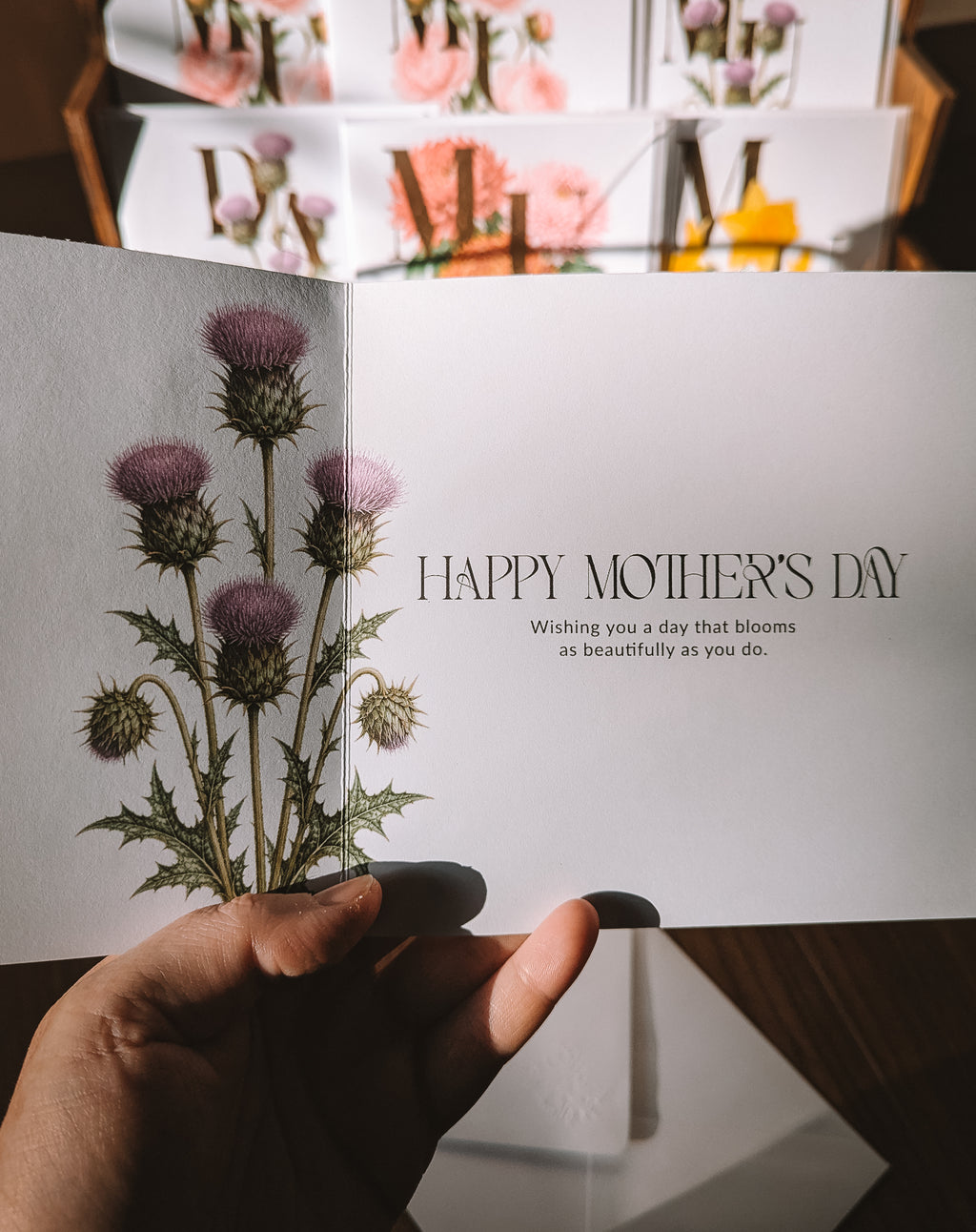 A hand holds an open greeting card with a botanical illustration of purple thistles on the left panel and the text 'HAPPY MOTHER'S DAY' on the right panel. The background shows more cards stacked in a box.