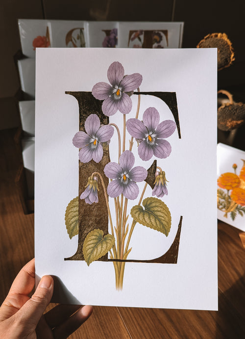 A hand holds a white art print featuring a large decorative letter E filled with purple violets and green leaves, set against a warm, blurred background of framed art and dried flowers.