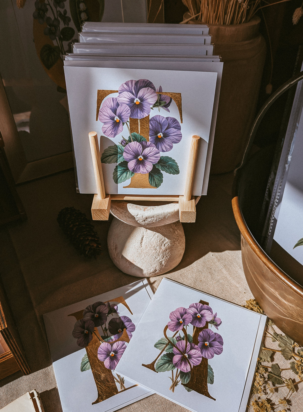 A stack of art print cards featuring purple violet flower illustrations displayed on a small wooden stand, with two additional cards laid out in front on a tabletop in a styled interior setting.