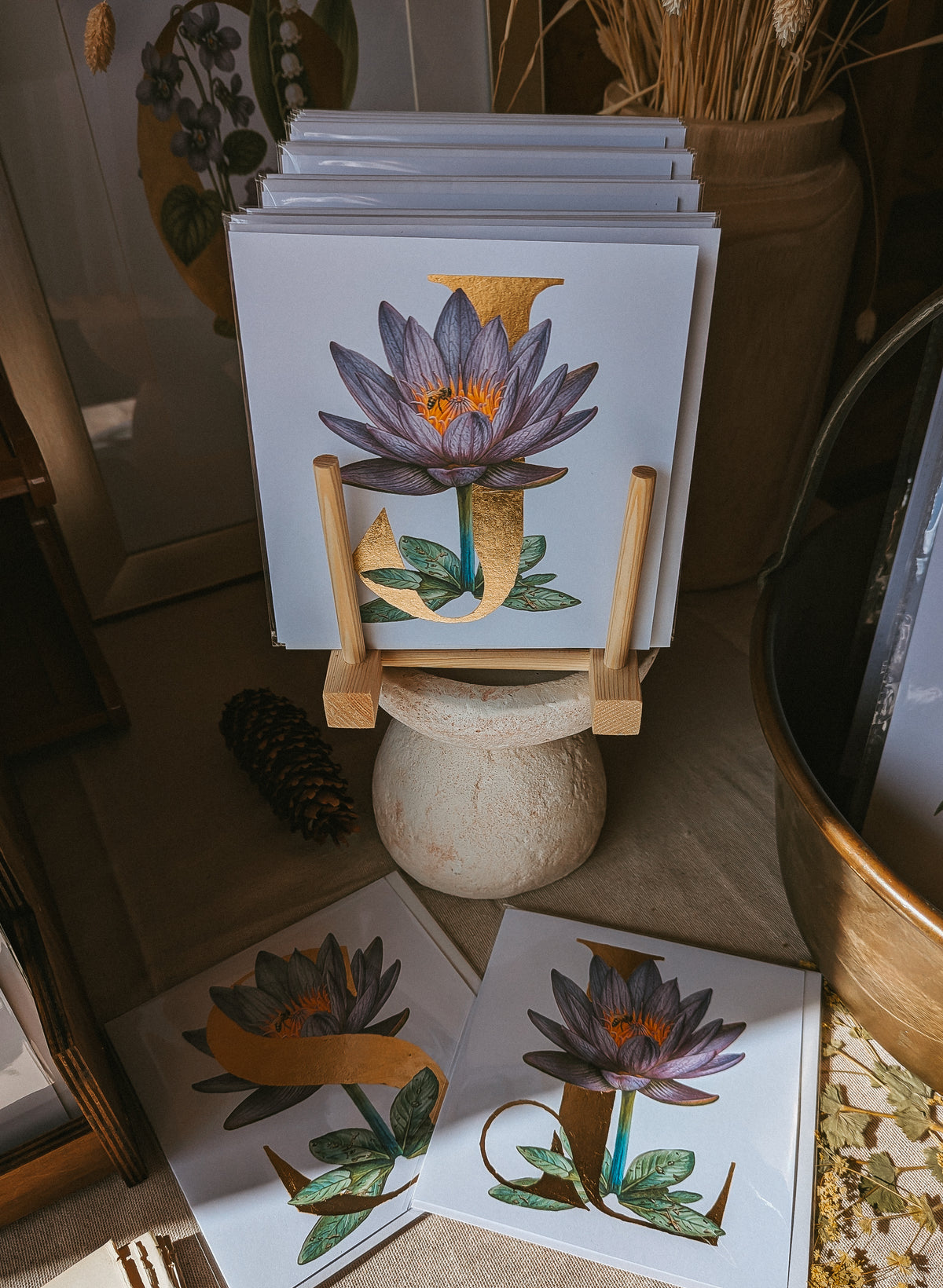 A stack of art cards featuring a purple water lily with gold and green botanical details, displayed on a small wooden stand in a rustic indoor setting with dried flowers and natural light.