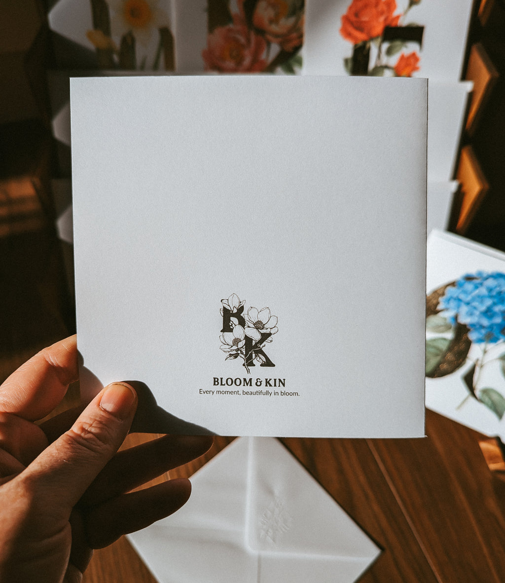 A hand holds a white square card printed with the Bloom & Kin logo and name, with floral artwork and additional cards visible in the background on a wooden surface.