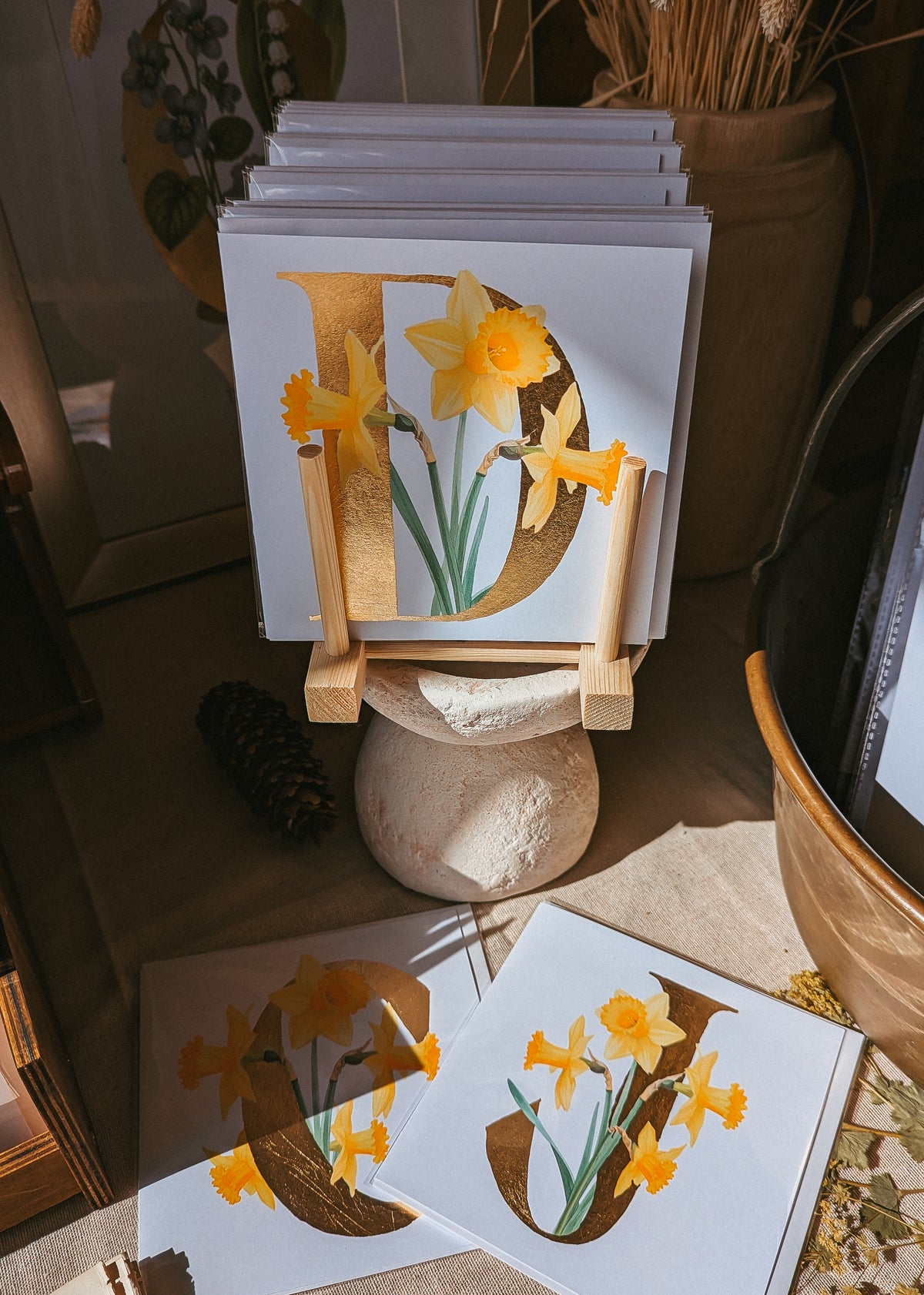 A stack of botanical art cards featuring the letter 'D' paired with a yellow daffodils, displayed on a small wooden stand in a styled interior setting.